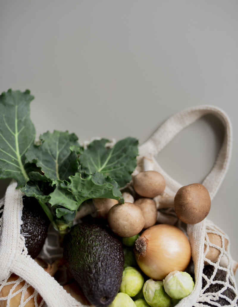 Cotton mesh bag filled with seasonal vegetables – kale, avocados, mushrooms, onion and Brussels sprouts – against a neutral background