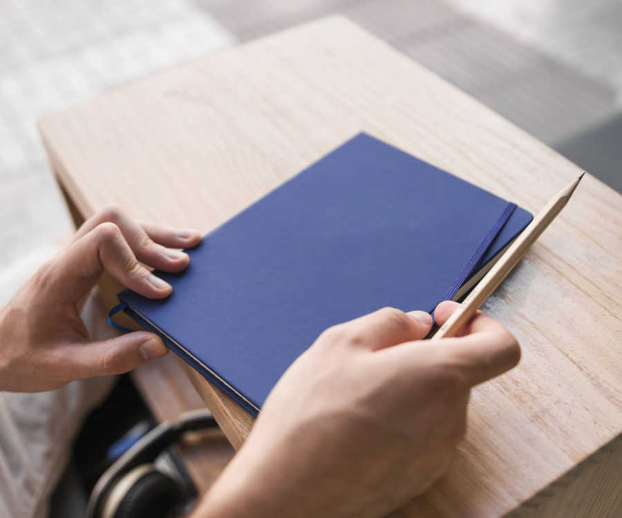 Hände halten ein blaues Notizbuch und einen Stift an einem Holztisch, Kopfhörer daneben — steht für persönliches, praxisnahes Coaching und Strategieplanung